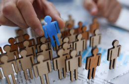 A businessman in blue shirt is holding a magnifying A businessman in blue shirt is holding object in his hand, is searching for personnel or people. Detective looking for missing person crowd of miniature figures choosing most suitable one